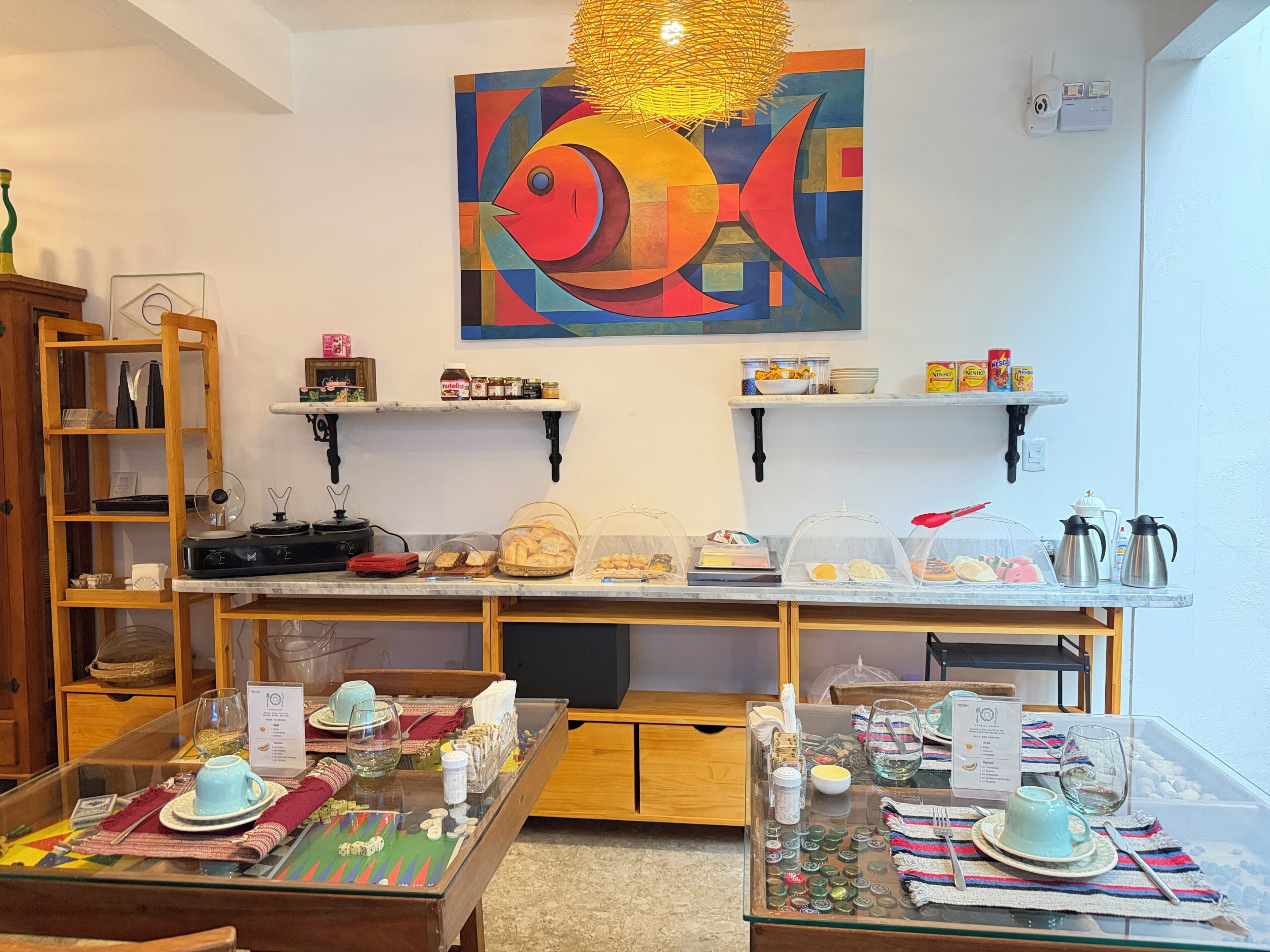 A dining area with tables set for breakfast, a fish painting on the wall, and a security camera.