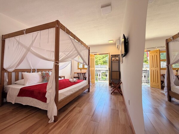 A four-poster bed with a canopy, a wooden floor, a small table with chairs, and a balcony with a railing.