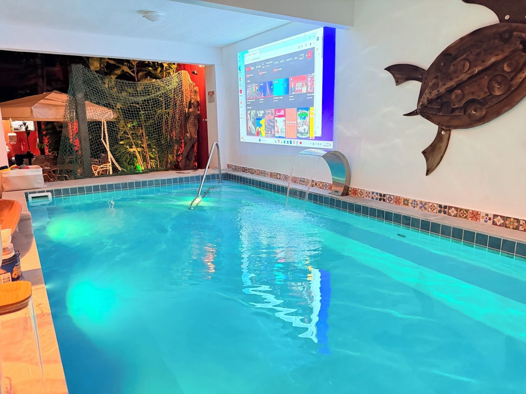 An indoor swimming pool with a tiled edge, a wall-mounted screen displaying a video, and a decorative fish sculpture.