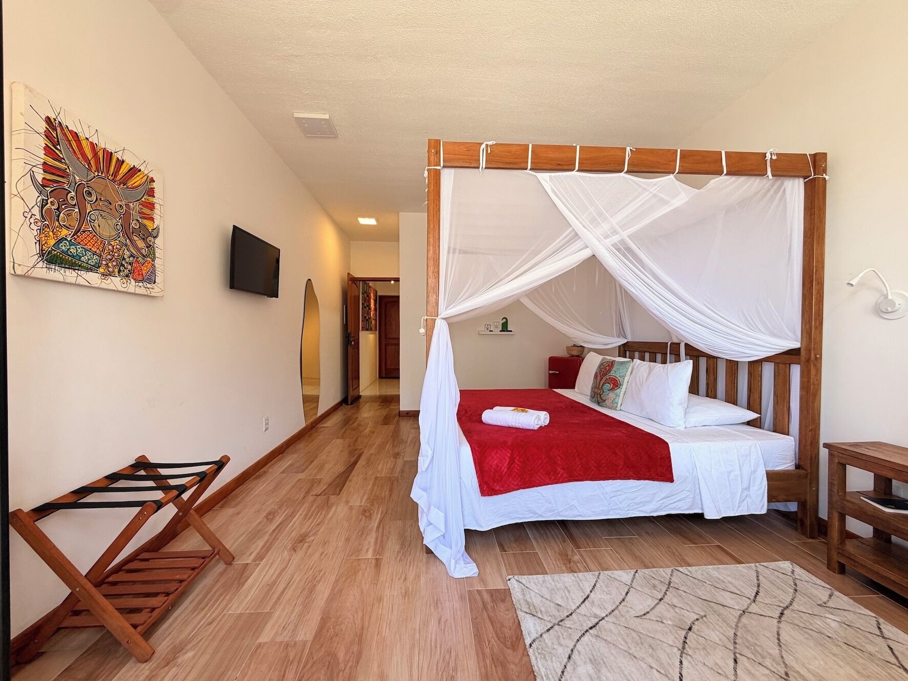 A bedroom with a four-poster bed, a wooden floor, a wall-mounted TV, and a colorful artwork.