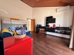 A compact living space with a kitchenette, a sofa, a red suitcase, and a wooden ceiling.