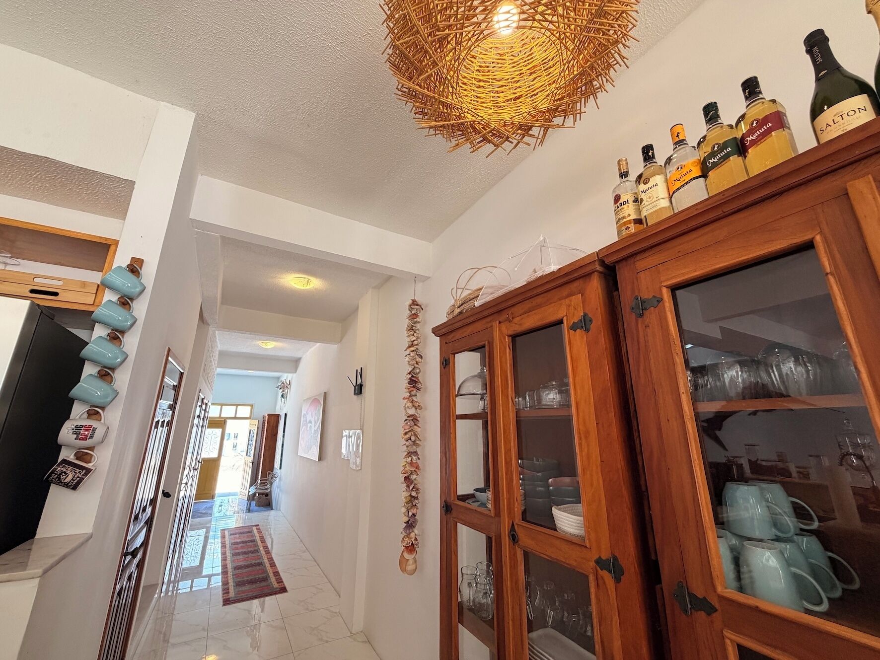 A hallway with a wooden cabinet containing glassware, a woven light fixture, and a row of blue mugs on the wall.