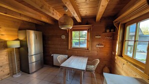 A wooden cabin interior with a kitchen, dining table, chairs, a refrigerator, a lamp, and a window with a view.