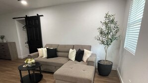 Ein Wohnzimmer mit einer Eckcouch, einem kleinen runden Tisch mit einer Vase, einer Topfpflanze und einer Schiebetür.
