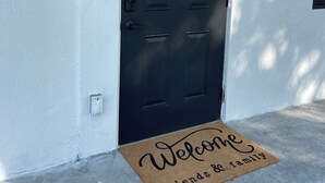 Eine schwarze Tür mit Fußmatte, auf der 'Willkommen Freunde & Familie' steht, und einem Lichtschalter an der Wand.