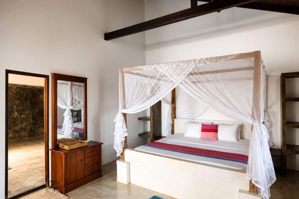 A bedroom with a four-poster bed, a wooden dresser, a mirror, and a view of the outside.