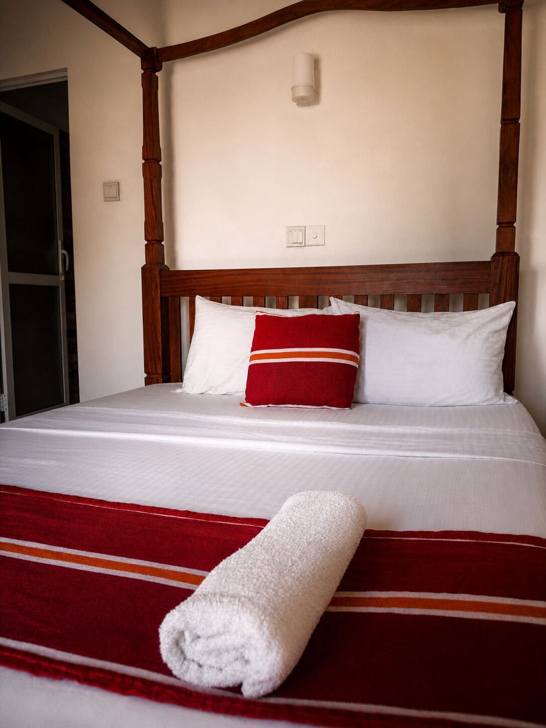 Un lit bien fait, recouvert d’une couverture à rayures rouges et blanches, avec une serviette blanche roulée et deux oreillers, un rouge et un blanc, dans une pièce dont la tête de lit est en bois et les murs sont blancs.