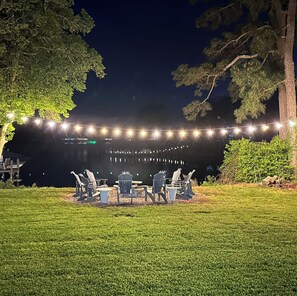 An outdoor seating area with chairs and tables, string lights overhead, and a grassy lawn.