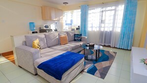 A living room with a sofa, a coffee table, a rug, a kitchen area with cabinets, and a window with curtains. - Peaceful 1-bedroom corporate apartment in vibrant Nairobi (Nairobi)