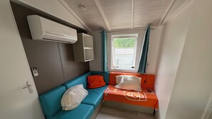Een kleine kamer met een blauwe bank, een oranje bed, een raam met gordijnen, en een airconditioning.
