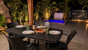 An outdoor dining area with a pool, surrounded by tropical plants and a wooden pergola.