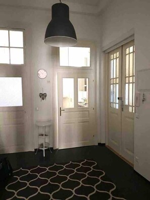 Un pasillo con un tapete estampado, una lámpara colgante negra, una puerta blanca con un espejo y una pared blanca con un reloj.