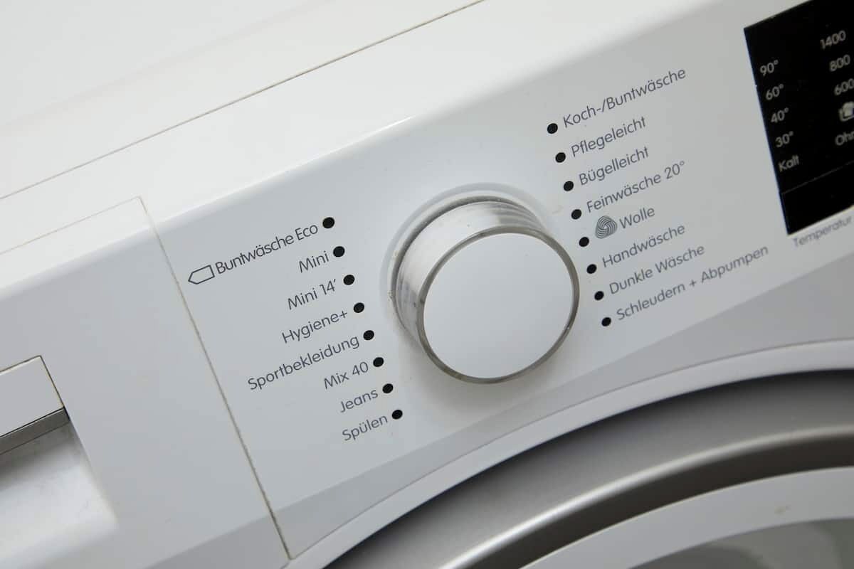 A washing machine with a control panel listing various wash cycles like 'Buntwäsche Eco,' 'Mini,' and 'Hygiene+.'