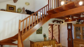 Een houten trap met een kroonluchter, een eettafel met stoelen en een houten kast met glazen deuren.