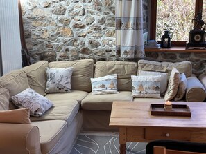 A cozy living area with a stone wall, a beige sofa with patterned cushions, a wooden table with a tray, and a window with curtains. - Quiet mountain house (ARIGNAC)