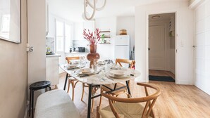 A dining area with a round table, chairs, a vase with red flowers, and a kitchen in the background.