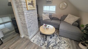 Ein Wohnzimmer mit grauem Ecksofa, einem runden Couchtisch, einem Teppich mit Marmormuster, einer Ziegelwand als Blickfang und einem dekorativen Wandbild.