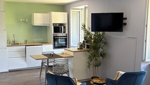 Een moderne keuken met witte kasten, een magnetron en een oven. Er is een eethoek met een tafel, stoelen en een aan de muur bevestigde televisie.