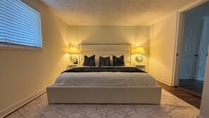 A bedroom with a large bed, two bedside lamps, a headboard, and a geometric-patterned floor. - Renovated 4BR, Minutes to Airport, Spacious & Cozy (Columbus)