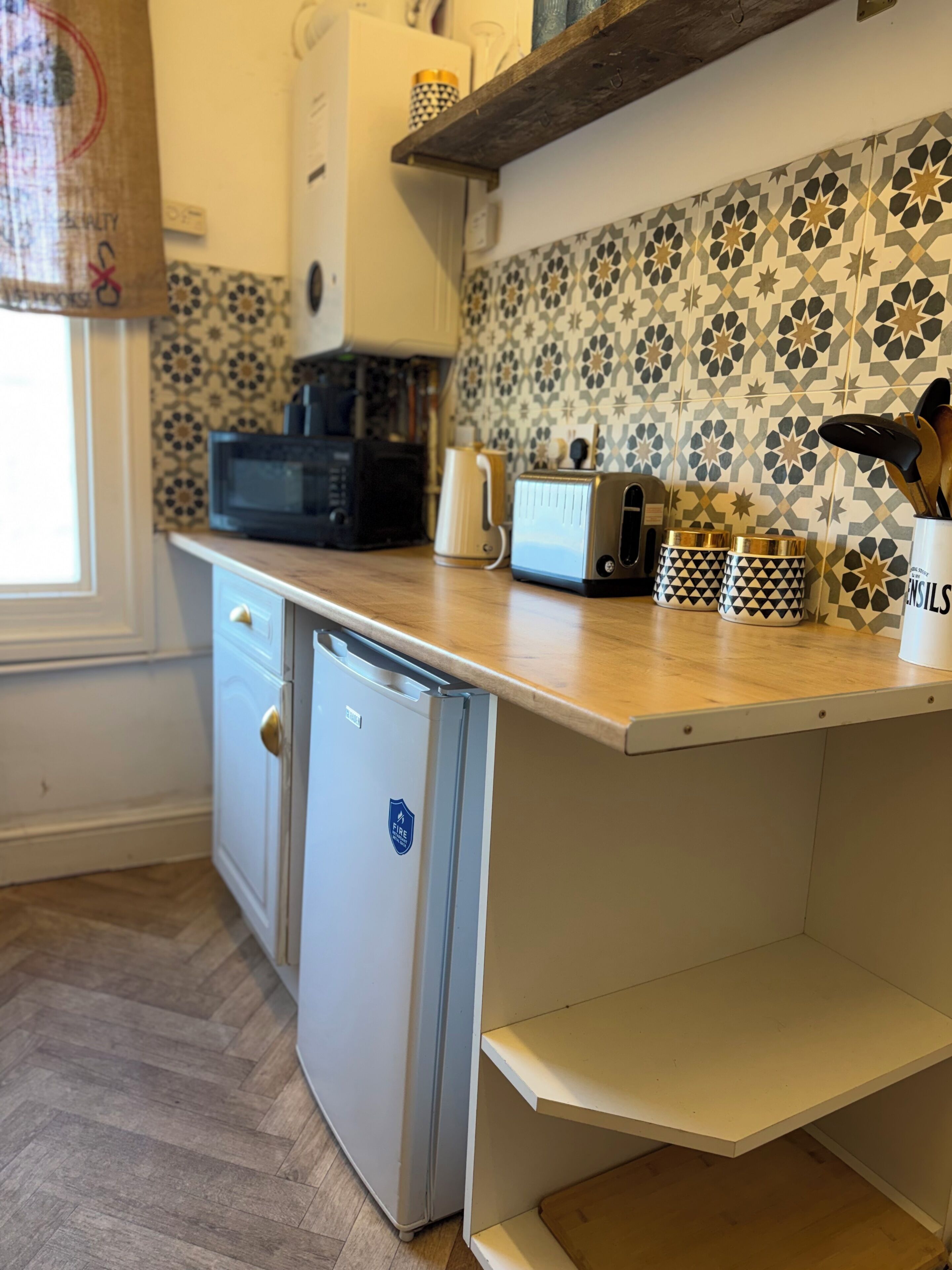 Ett kompakt kök med en mikrovågsugn, brödrost och kaffebryggare på bänkskivan, ett litet kylskåp med en klistermärke som visar brandsäkerhetsinformation och mönstrade kakelplattor ovanför arbetsytan.