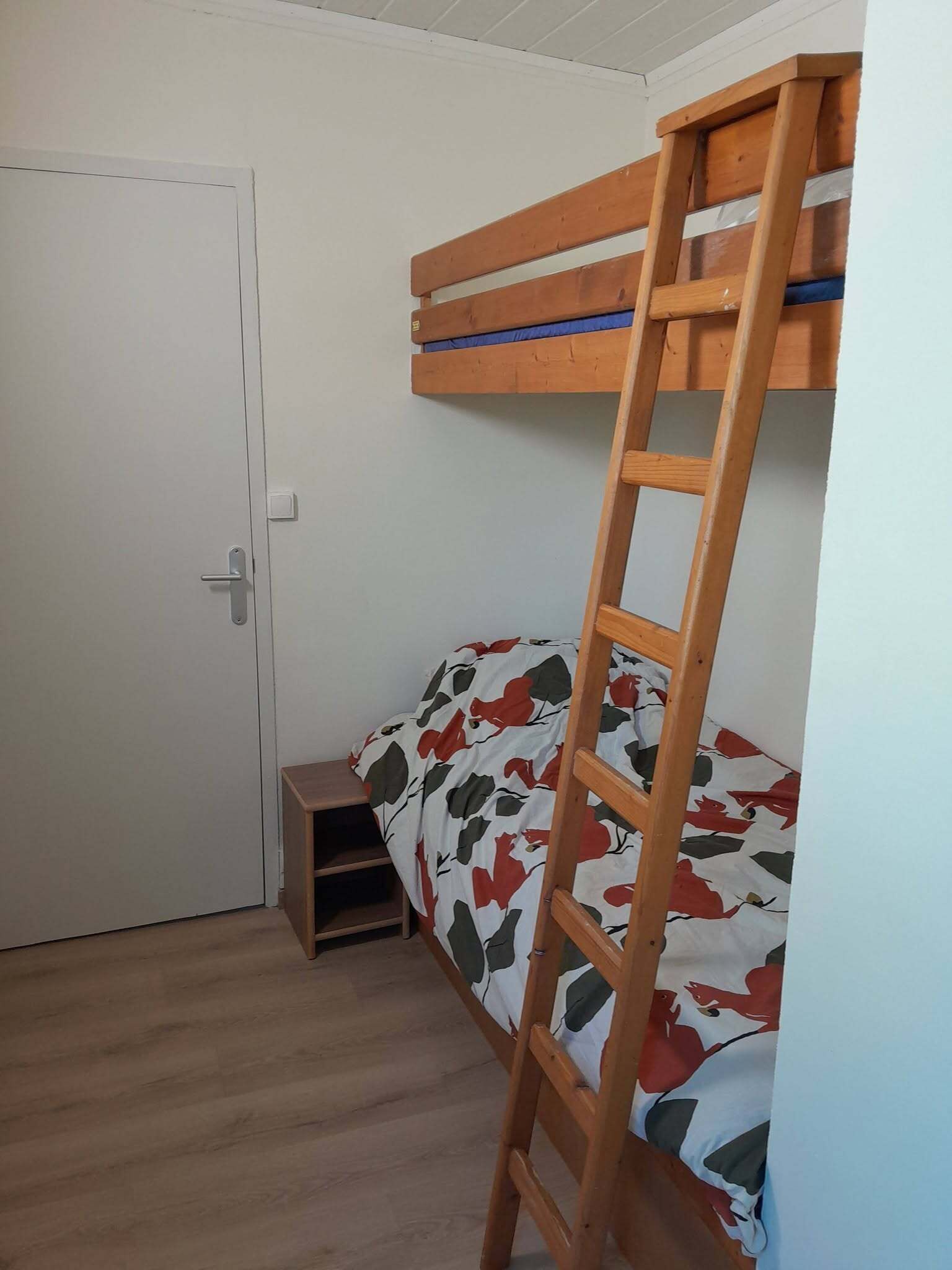 Un lit superposé avec une échelle en bois, un petit tabouret en bois et une porte dans une pièce dont le sol est recouvert de planches de bois.