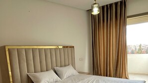 Ein Hotelzimmer mit einem großen Bett, einem Kopfteil, einem Nachttisch, einer Stehlampe, einem Fenster mit Vorhängen und Blick auf Gebäude.