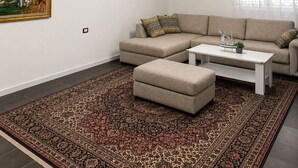A living room with a sectional sofa, ottoman, patterned rug, wooden ceiling beams, pendant lights, a chandelier, and a framed painting.