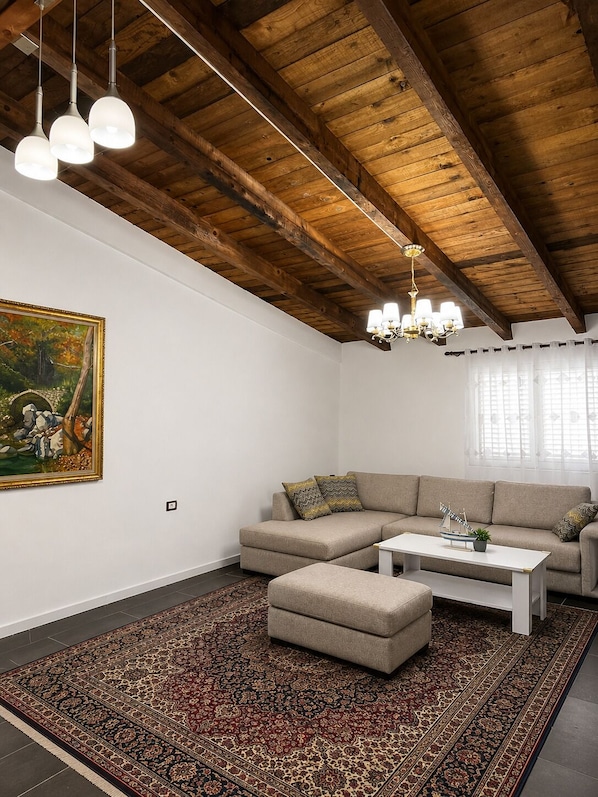 A living room with a sectional sofa, ottoman, patterned rug, wooden ceiling beams, pendant lights, a chandelier, and a framed painting.