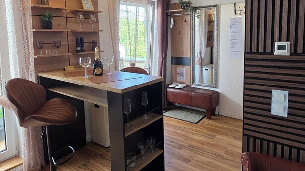A modern living room with a wooden floor, a brown leather sofa, a black bar stool, a wooden dining table with wine glasses, and a pendant light. - Apartment with high-quality furnishings and underground parking space in Burghausen. (Burghausen)
