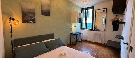 A bedroom with a bed, two wall-mounted light fixtures, a window with a view, a radiator, and a mirror.
