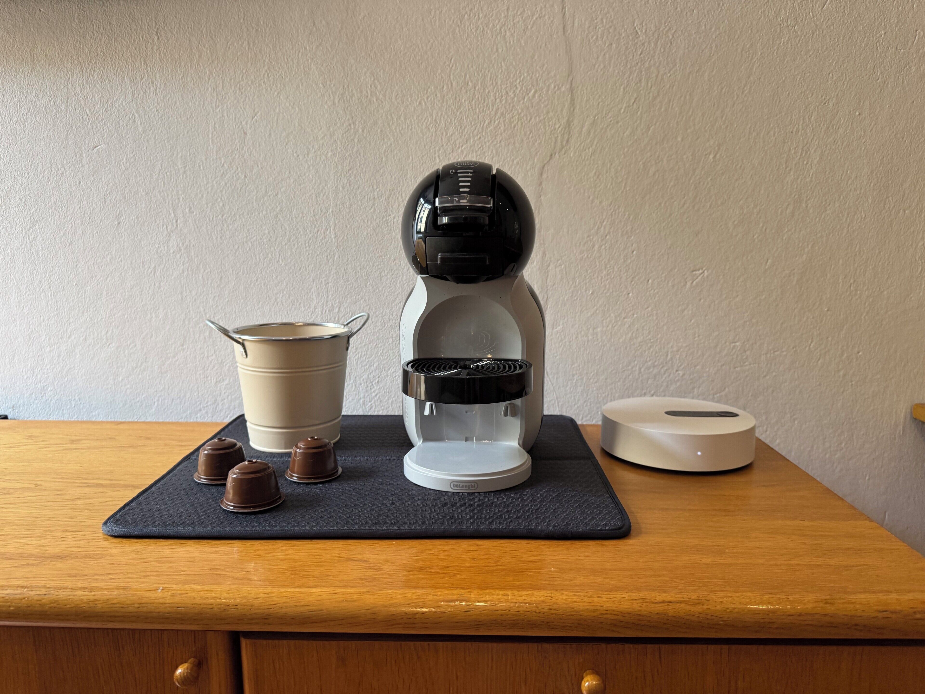 Une machine à café est posée sur une table en bois, accompagnée d’un tapis, d’un récipient et d’un rouleau d’essuie-tout.