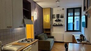 Eine kleine Küche mit Gasherd, Backofen und Spüle. Ein Sofa und ein Esstisch mit Stühlen sind ebenfalls vorhanden. Das Zimmer verfügt über ein Fenster mit Jalousien und eine Heizung.
