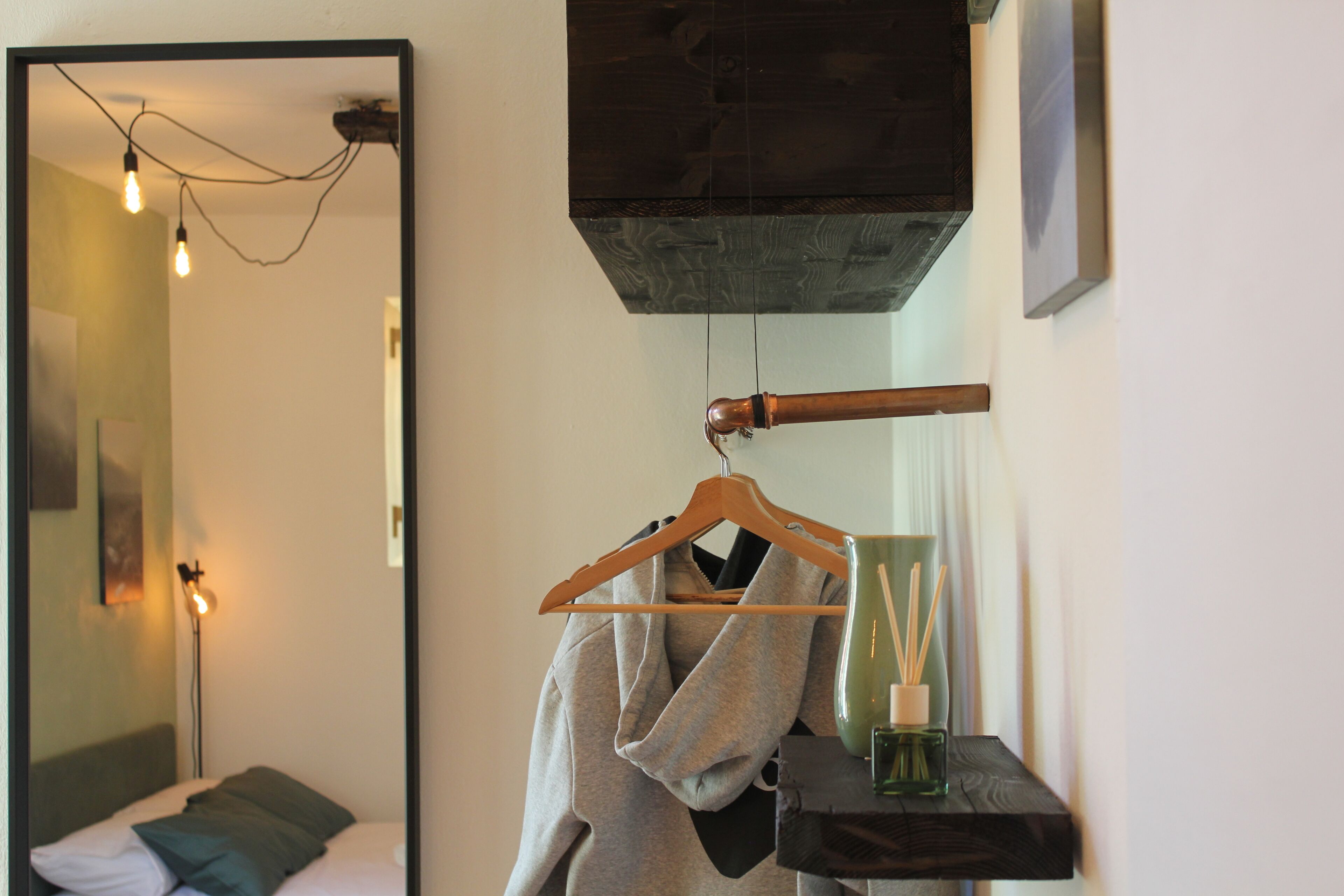 Une chambre d’hôtel avec un lit, un peignoir suspendu, un cintre en bois, un vase vert et une petite étagère.