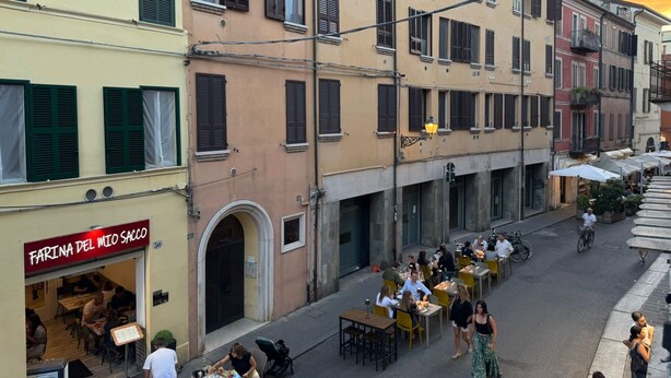 Eine Straße mit Außenbestuhlung, ein Restaurant namens 'Farina del Mio Sacco' und mehrstöckige Gebäude mit Balkonen.