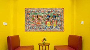 A room with two red leather chairs, a small wooden table with a vase of flowers, and a colorful tapestry on the yellow wall. - Hotel Ram Janaki (Janakpur)
