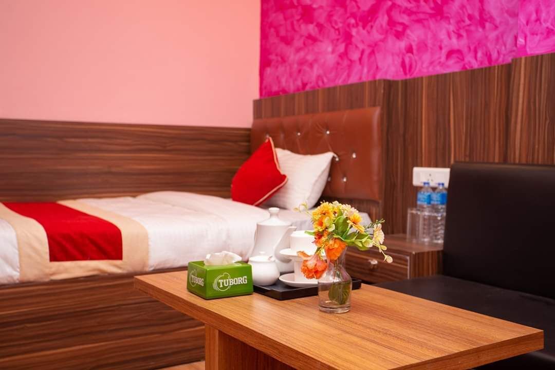 A hotel room with a bed, a wooden nightstand with a vase of flowers, a box of tissues, and a water bottle on the wall.