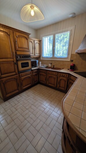 Une cuisine avec des meubles en bois, un four encastré, un four à micro-ondes, un évier et une fenêtre donnant sur les arbres.