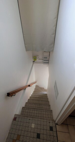 Un escalier avec une rampe, un sol carrelé et un mur blanc percé d’une grille d’aération.