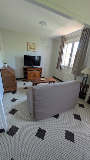 Ein Wohnzimmer mit einer Couch, einem Fernseher, einem Holzschrank, einem gemusterten Boden und einem Fenster mit Vorhängen.