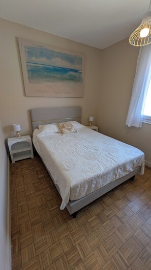 Ein Schlafzimmer mit einem Bett, Nachttischen, einer Lampe, einem Gemälde und einem Fenster mit Vorhängen.