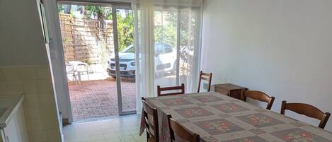 Un espace repas comprenant une table, des chaises, un luminaire au plafond et une vue sur l’extérieur, accessible par une porte vitrée.