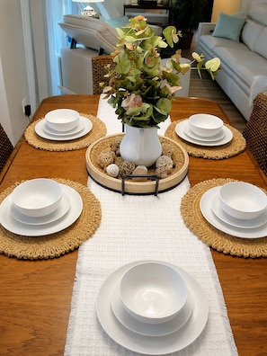 Mesa de jantar posta com pratos e tigelas brancos, um vaso branco com flores e objetos decorativos.