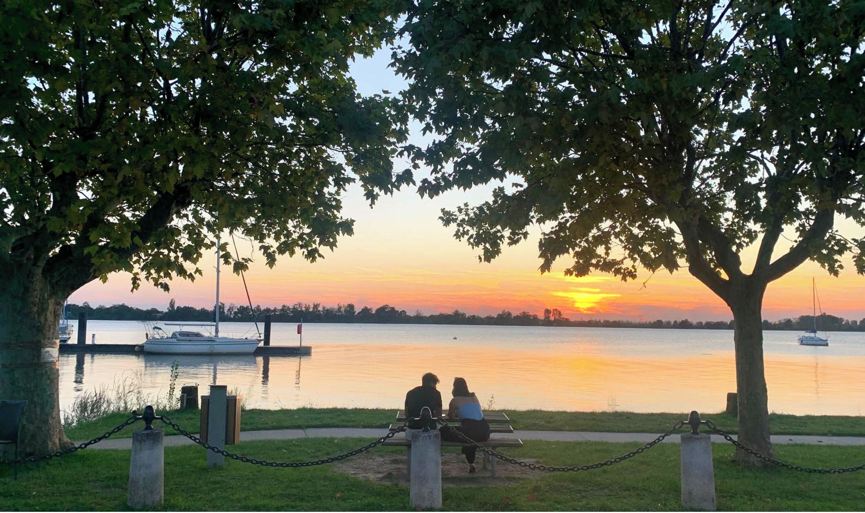 Vista del atardecer desde un banco en un parque, con un lago tranquilo y barcos.