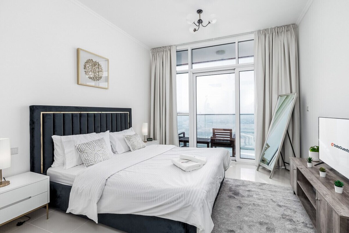 Ein modernes Hotelzimmer mit einem großen Bett, einem Nachttisch, einem Tischchen, einem Fernseher, einem Fenster mit Vorhängen und Blick auf die Stadt.
