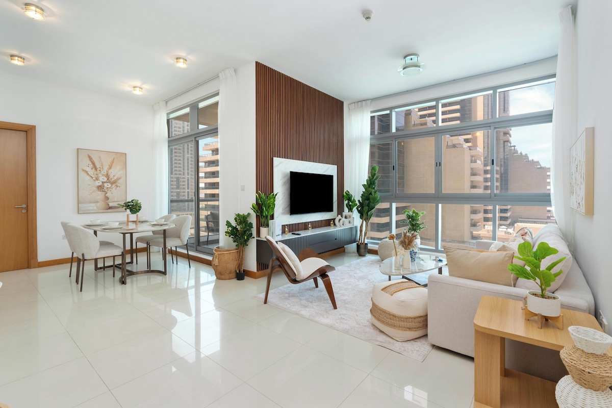 A modern living room with a flat-screen TV, dining area, and large windows offering city views.