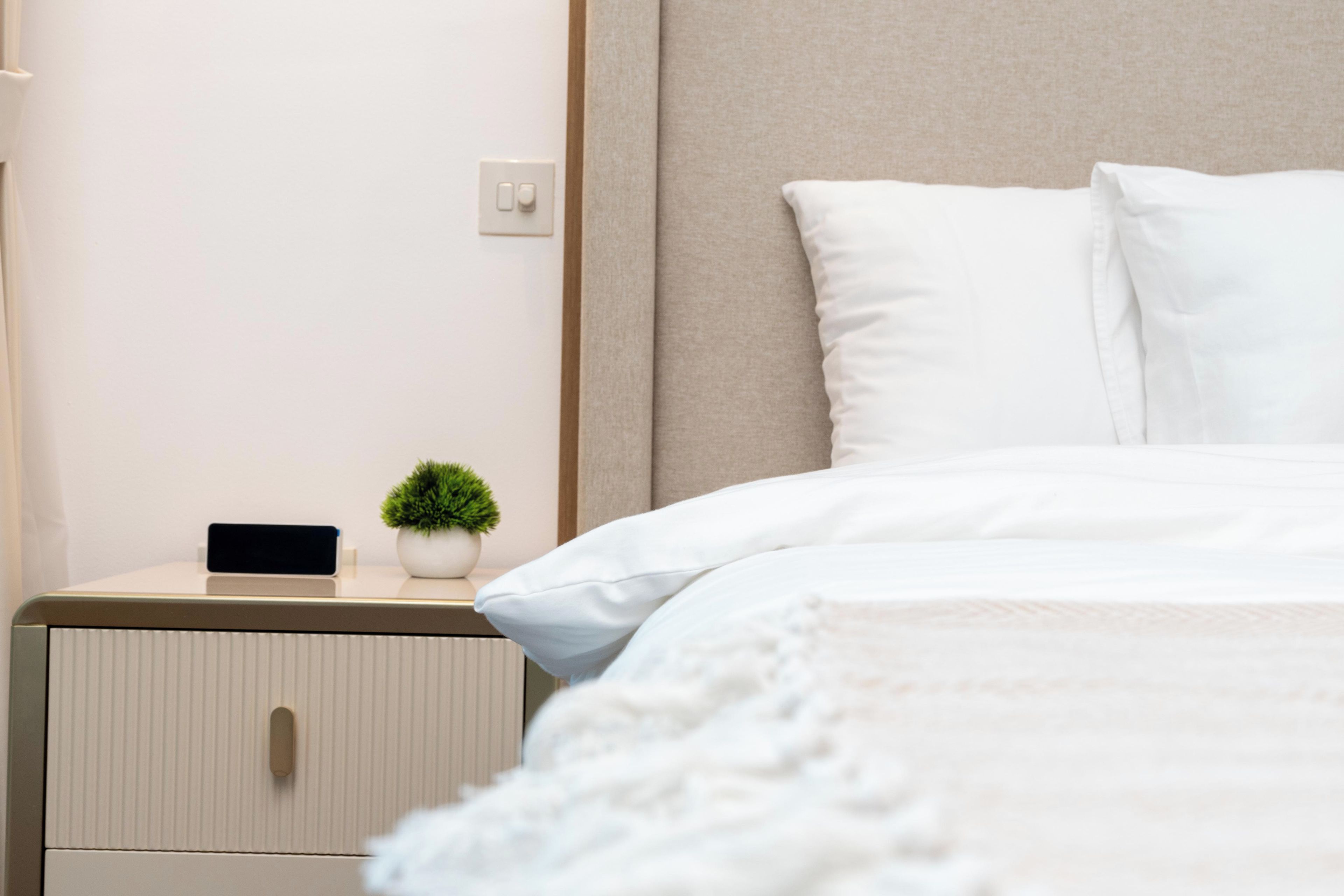 Ein ordentlich bezogenes Bett mit weißen Leinentüchern, ein kleiner Nachttisch mit einer Pflanze und ein an der Wand befestigter Lichtschalter.