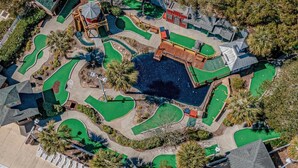 An aerial view of a golf course with multiple green areas, a central water feature, and surrounding trees. - Steps to Sand, Ocean Views, Water Park & Golf Cart (Myrtle Beach)