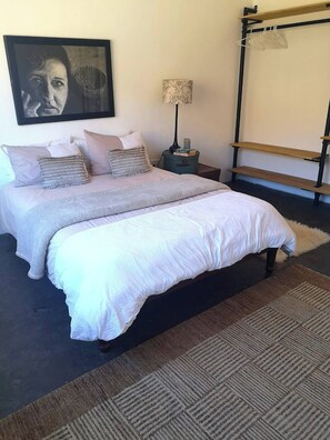 A bedroom with a bed, pillows, a nightstand, a lamp, a hanging rack, and a framed picture on the wall. - Tranquil Karoo Retreat (Laingsburg)