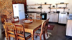 A dining area with a wooden table and chairs, a refrigerator, a stove, and shelves with kitchen items. - Tranquil Karoo Retreat (Laingsburg)