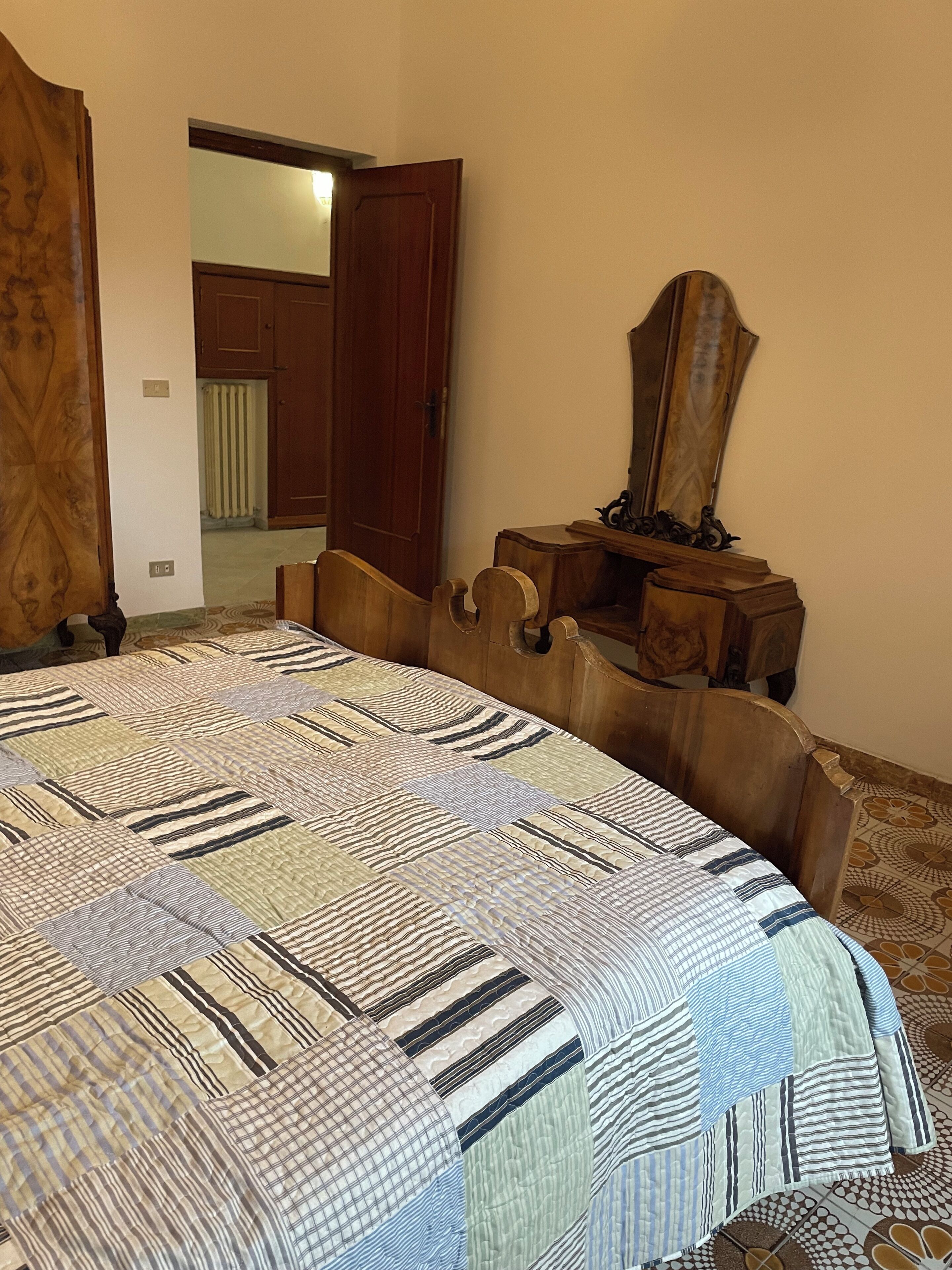 A bedroom with a wooden headboard, a large bed with a patterned quilt, a wooden dresser with a mirror, and an open door leading to another room.